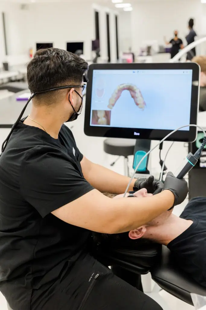 A dental professional uses a digital scanner on a patient, with an image of teeth displayed on a screen in a modern dental clinic.