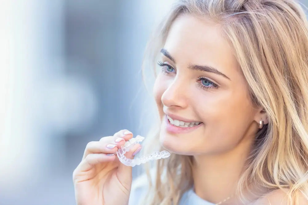 A woman with blonde hair holds a clear dental aligner near her mouth, smiling. She is outdoors with a blurred background, contemplating the Invisalign cost in Tallahassee, FL.