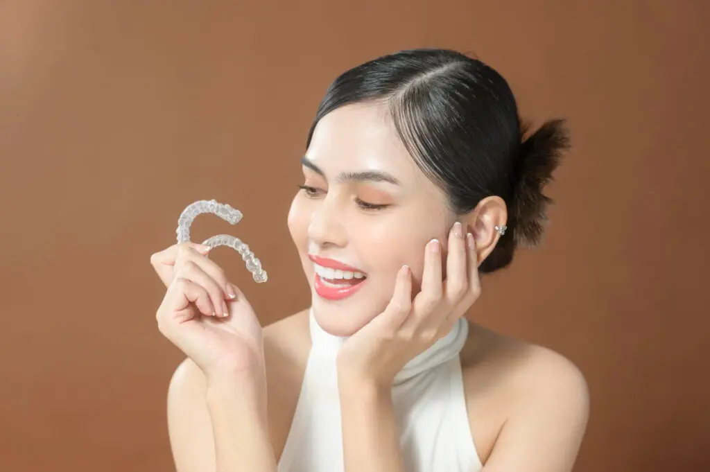Person smiling while holding clear dental aligners, against a brown background at Redemption orthodontics in Tallahassee