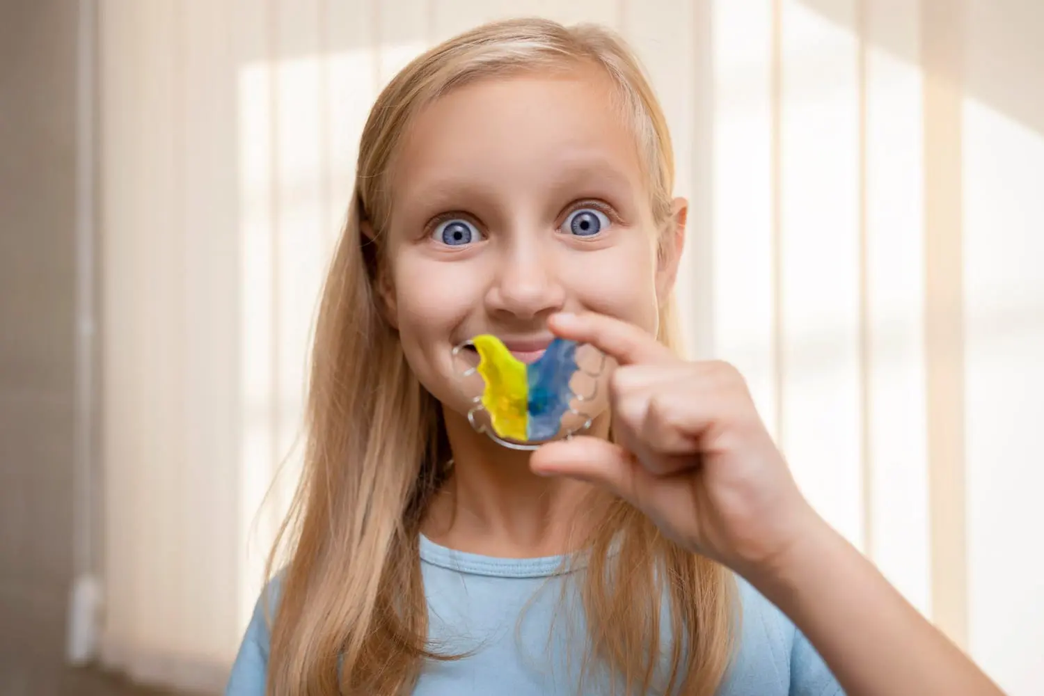 Girl holding a colorful dental retainer, wearing a light blue shirt, standing in front of vertical blinds at Redemption orthodontics in Tallahassee.