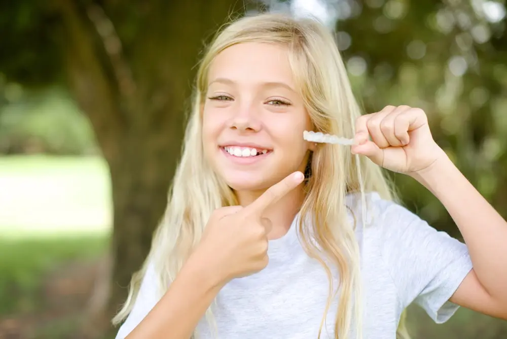 A young girl beams with joy while holding and pointing to her Invisalign clear aligners. She stands outdoors, with the gentle rustle of trees in the background, perfectly capturing a moment of carefree happiness in Tallahassee, FL