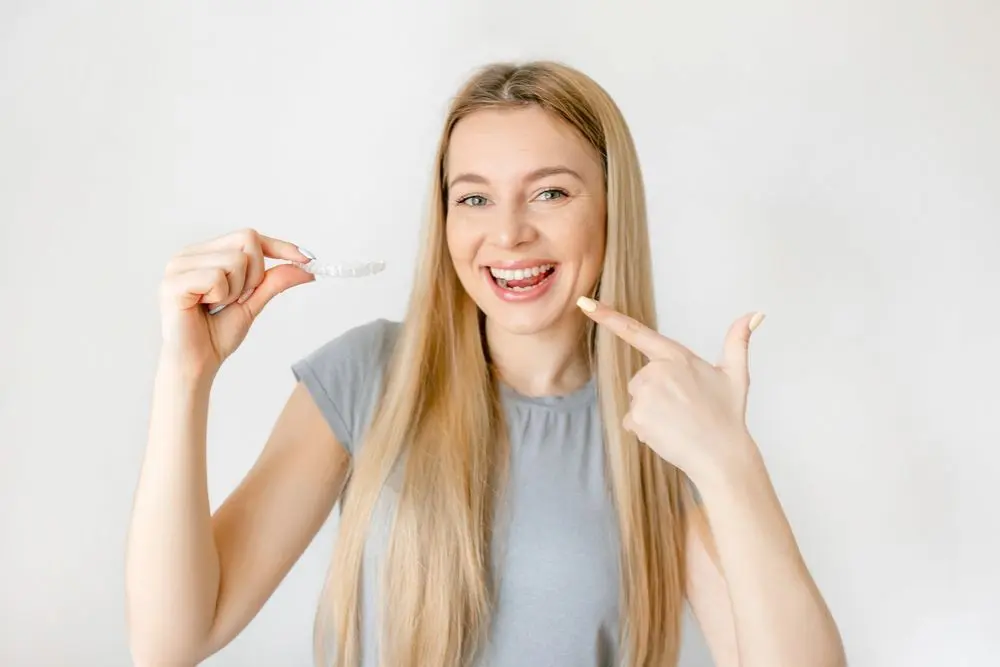 Woman smiling and pointing to her teeth, holding a clear dental aligner in her other hand, showcasing Adult braces from Redemption Orthodontics in Tallahassee, FL.