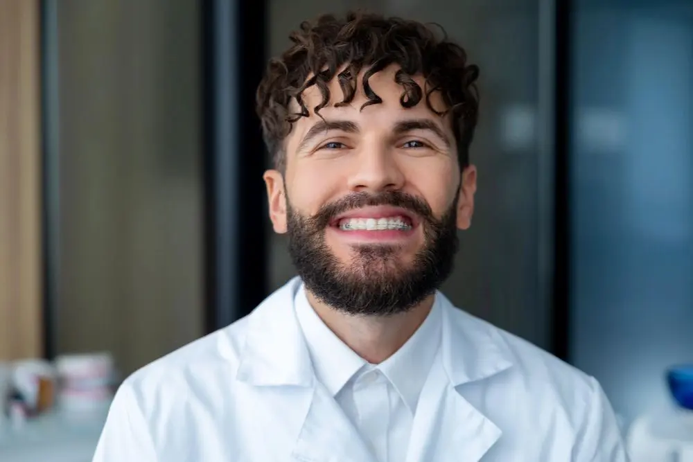 A person with a beard and curly hair smiles while wearing a white coat showing his adult ceramic braces from Redemption Orthodontics in Tallahassee, FL.