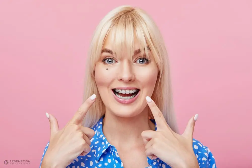 Smiling person with braces, wearing a blue polka dot shirt, pointing at their metal braces as one of the alternatives to braces in tallahassee, fl against a pink background.
