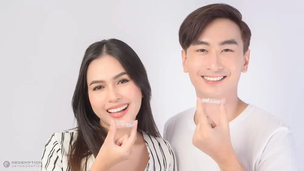 A woman and a man smiling and holding clear dental aligners in their hands as an example to show how to fix overbite in Tallahassee, FL against a plain white background.