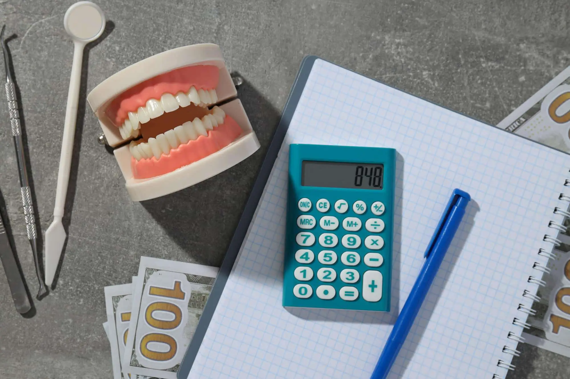 Dental model, calculator, blue pen, and dental tools on a notepad, with US hundred-dollar bills partially visible underneath. representing to cheap invisalign costs at Redemption Orthodontics in Tallahassee, FL.