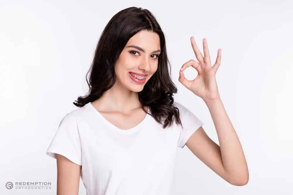 Young woman with long dark hair and braces smiles, wearing a white t-shirt and making an "OK" hand gesture against a plain white background after being explained about "How Long Do Braces Take in Tallahassee, FL".