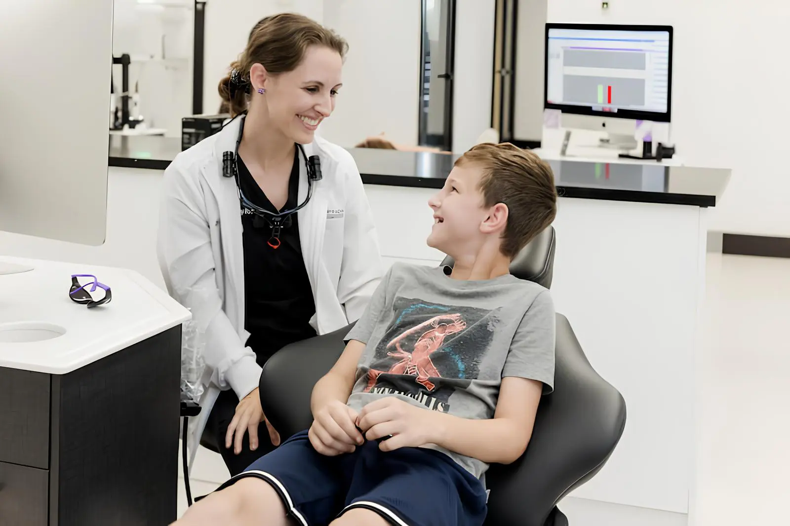 A dentist talks with a smiling boy sitting in a dental chair about invisalign cost, with a computer and dental equipment in the background in Tallahassee, FL.
