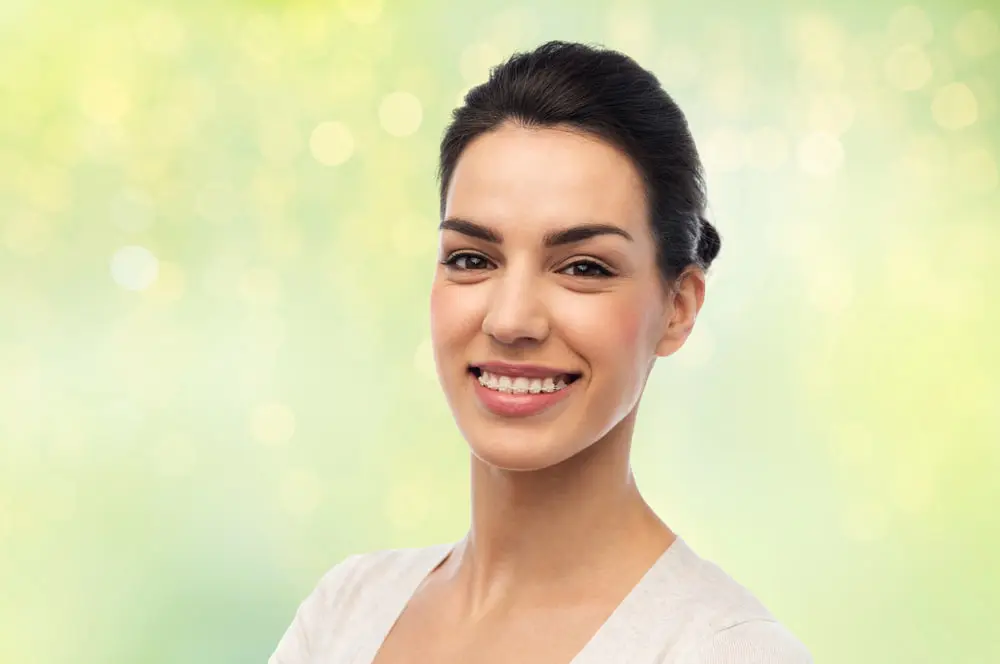 A woman with dark hair tied back smiles confidently at the camera, showing her adult braces in a blurred green and yellow outdoor setting, representing Redemption Orthodontics in Tallahassee, FL.