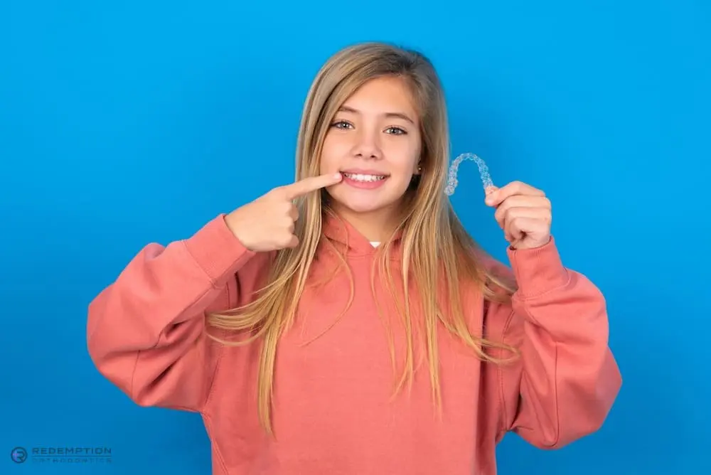 A girl in a pink hoodie smiles and points to her teeth while holding a clear aligner after understanding Does Insurance Cover Invisalign in Tallahassee and Bradfordville, FL