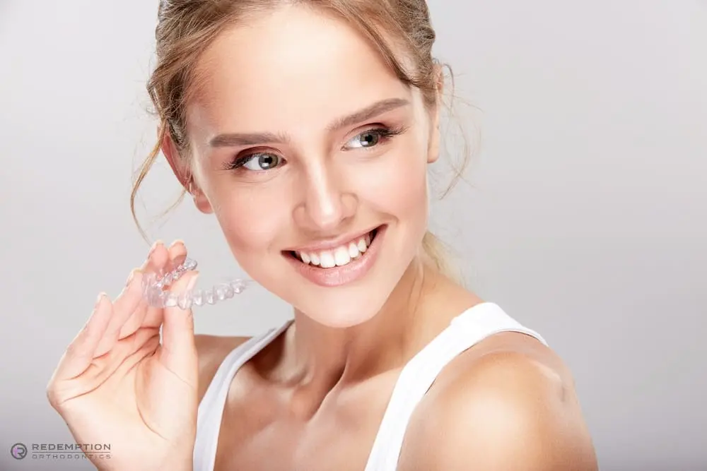 A woman in a white tank top smiles, holding a clear dental aligner near her face after understanding Does Insurance Cover Invisalign in Tallahassee and Bradfordville, FL
