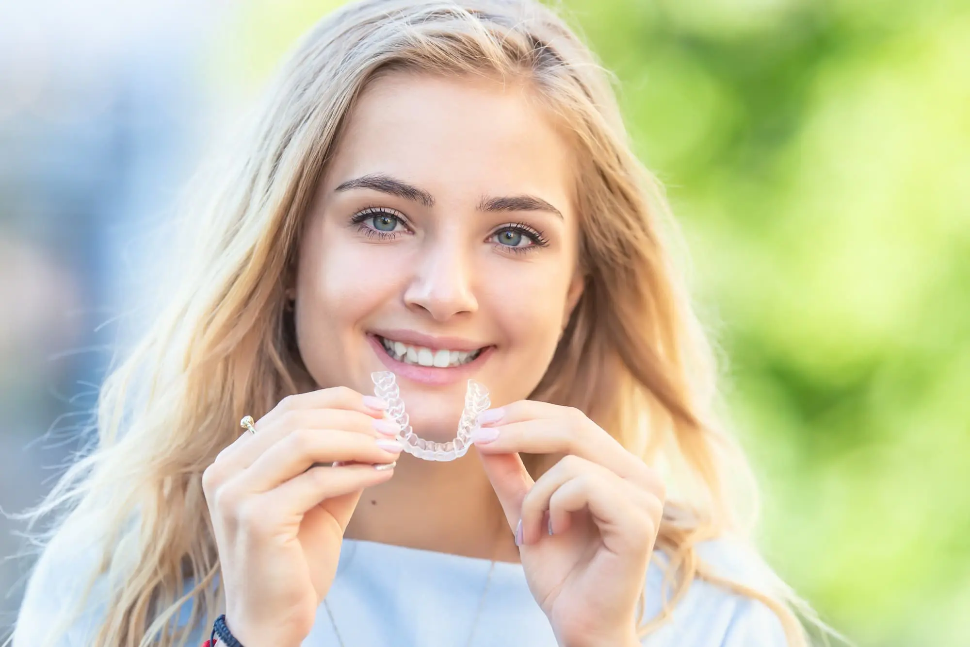 Outdoors in Tallahassee, FL, a person with long blonde hair smiles while holding a clear dental aligner from Redemption Orthodontics, showcasing an Invisalign affordable treatment option.