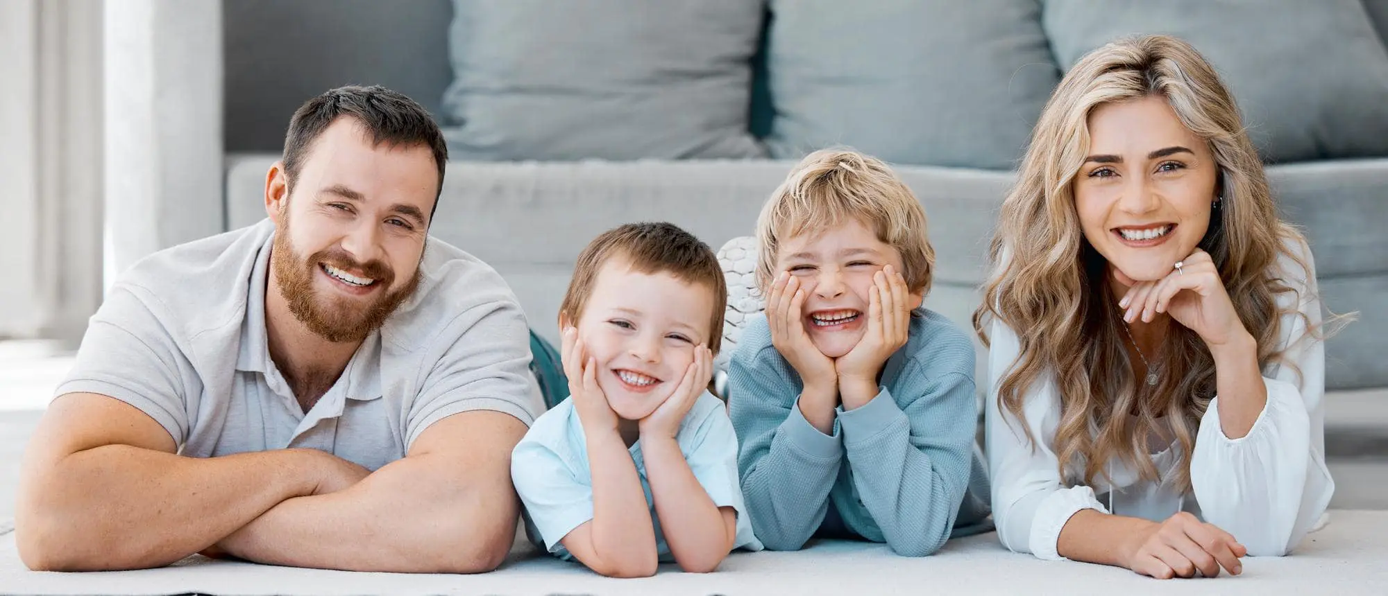 A man, woman, and two young boys lie indoors with their heads propped on their hands, smiling at the camera—a joyful moment captured at Redemption Orthodontics in Tallahassee, FL, highlighting Affordable Invisalign options for families.