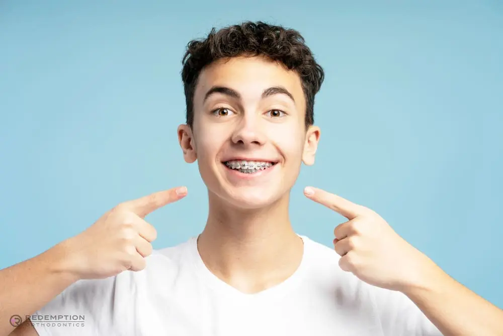 A teenage boy with curly hair smiles and points at his dental braces against a light blue background, representing Redemption Orthodontics' braces for overbite in Tallahassee, FL.