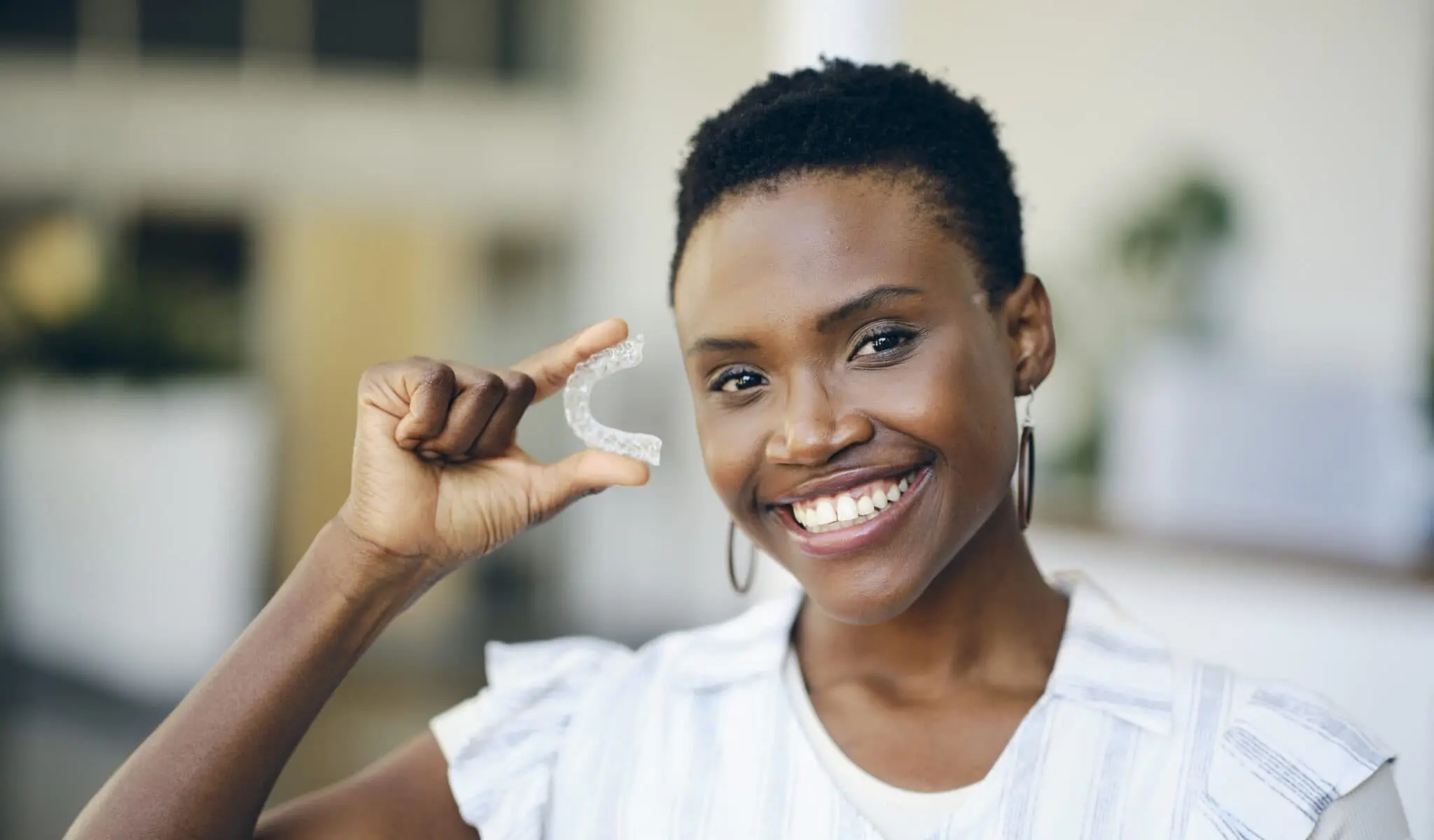 A woman at Redemption Orthodontics in Tallahassee, FL smiles indoors, holding a clear dental aligner in her right hand against a blurred background, highlighting how Invisalign can be covered by insurance.