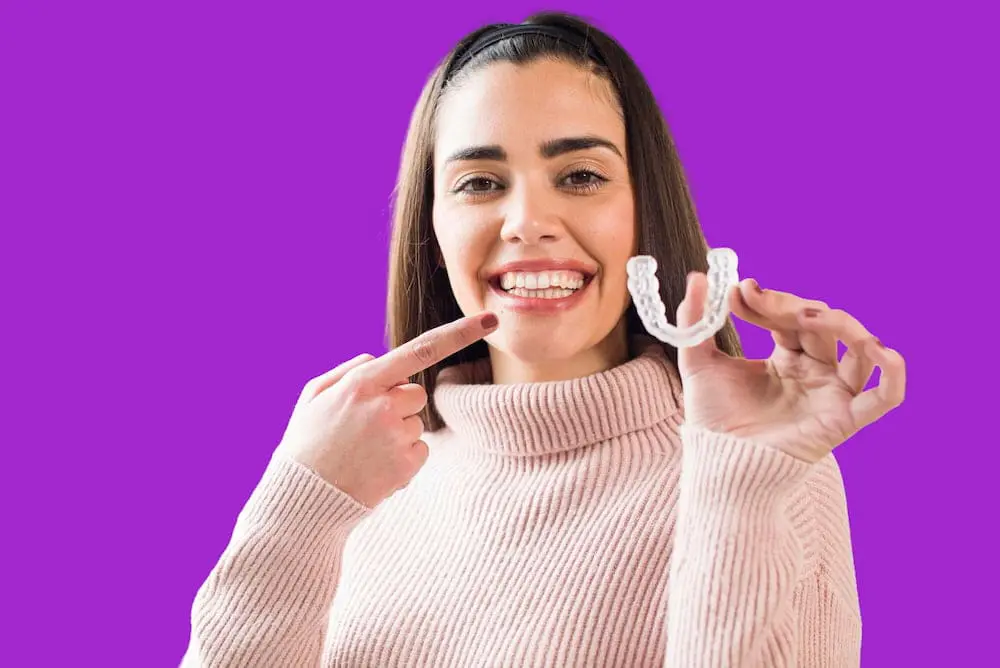 A woman in a light pink sweater smiles and points at her teeth while holding a clear dental aligner, set against a solid purple background, representing invisalign dental insurance at Redemption Orthodontics in Tallahassee, FL.