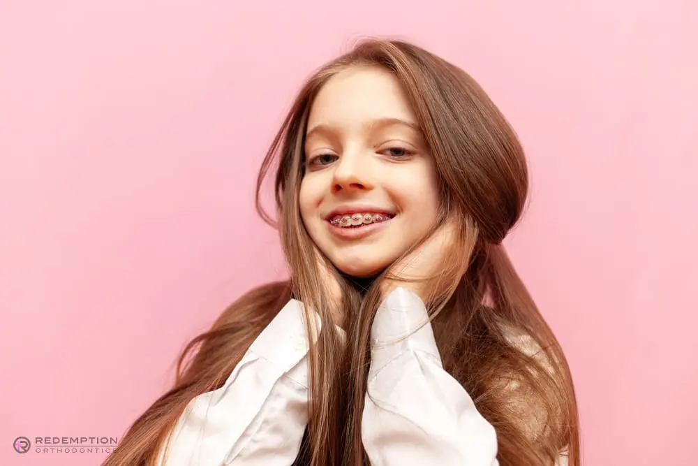A young girl with long brown hair and braces smiles, holding her face with both hands, set against a pink background at Redemption Orthodontics' braces for overbite in Tallahassee, FL.