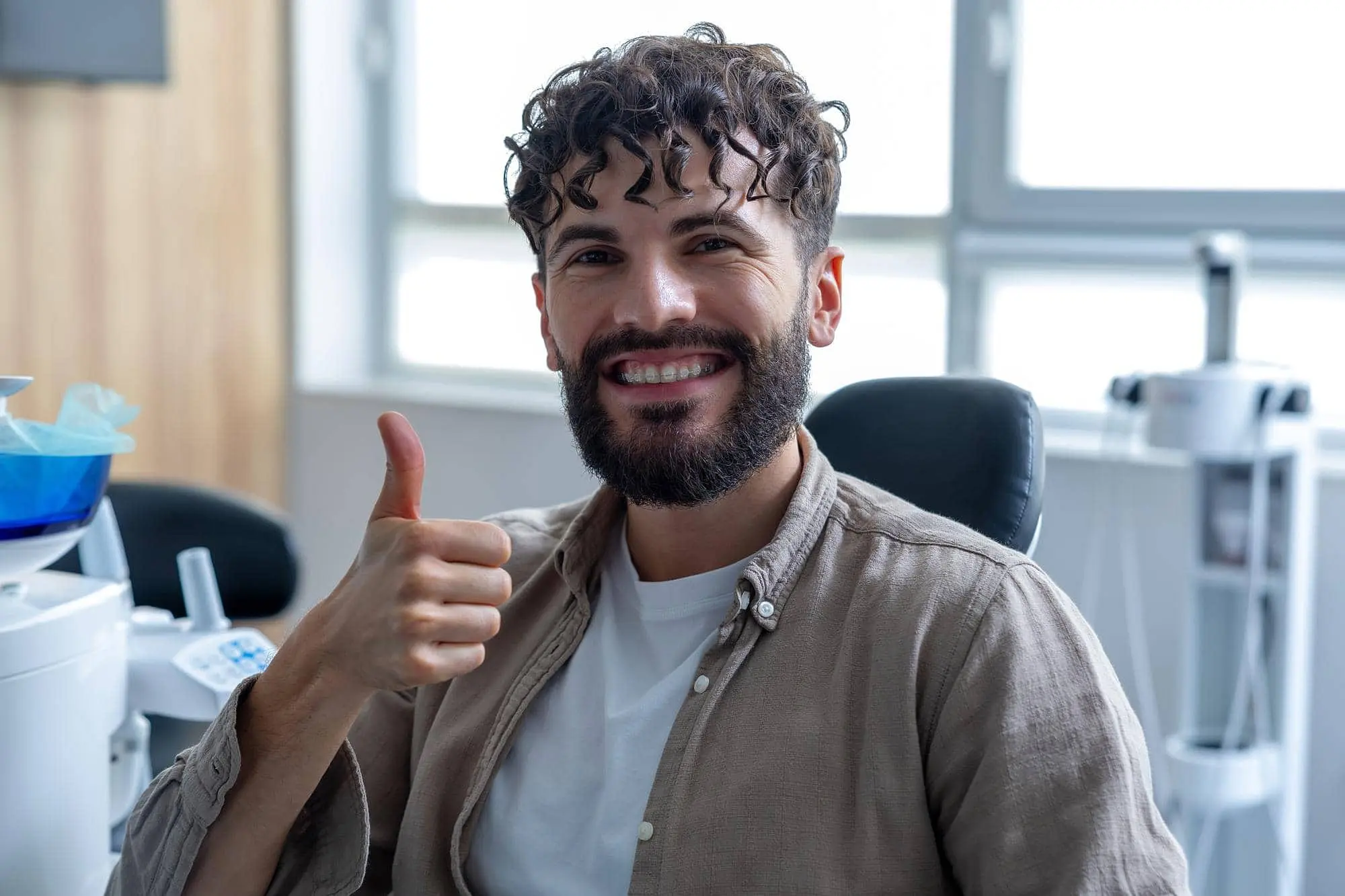 A bearded man with curly hair smiles with ceramic braces and gives a thumbs up at Redemption Orthodontics in Tallahassee, FL.