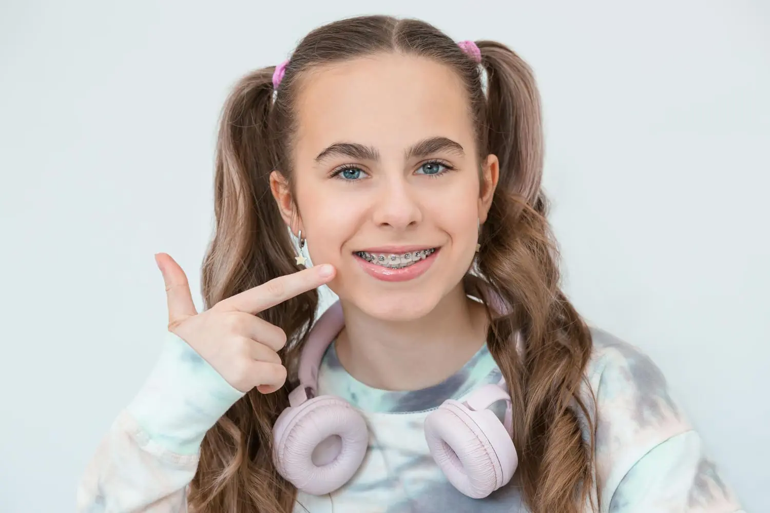 Smiling teen with pigtails and metal braces points to her teeth, wearing headphones; Redemption Orthodontics in Tallahassee, FL.