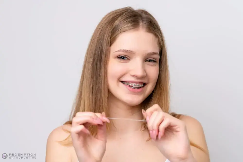 Smiling teen girl with long blonde hair and braces holds dental floss, representing Redemption Orthodontics in Tallahassee, FL - How to Floss with Braces in Tallahassee, FL