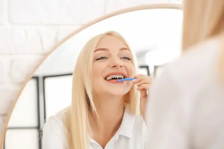 Smiling, a blonde woman brushes her teeth in front of a mirror at Redemption Orthodontics in Tallahassee, FL - How to Floss with Braces in Tallahassee, FL