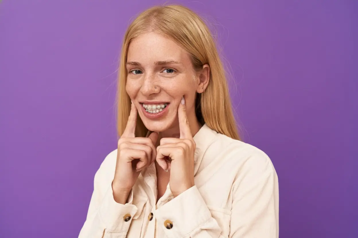 A smiling young woman with blonde hair points to her ceramic braces at Redemption Orthodontics in Tallahassee, FL.