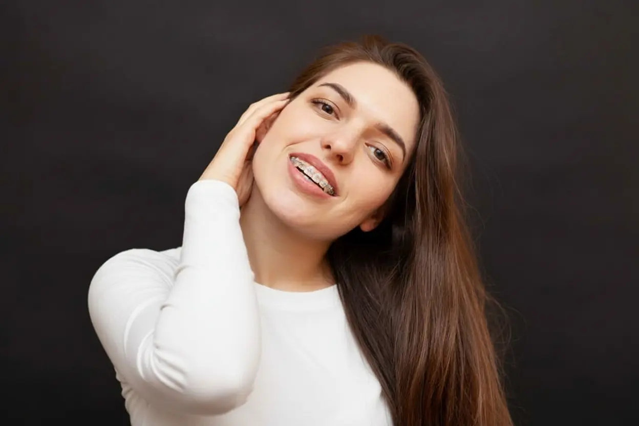 Smiling woman with ceramic braces stands against a yellow background at Redemption Orthodontics in Tallahassee, FL, touching her neck.