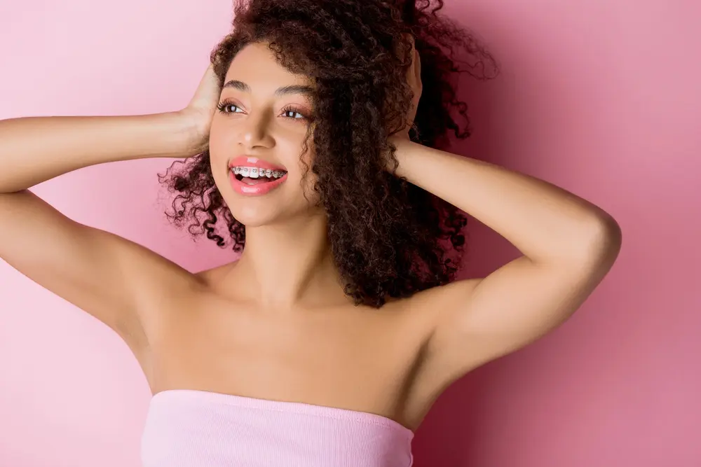 A smiling woman with curly hair and braces poses in front of a pink background at Redemption Orthodontics in Tallahassee, FL - Clear Braces vs Metal Braces in Tallahassee, FL