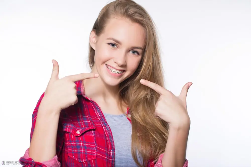 Teen girl in a red plaid shirt points at braces, smiling; Redemption Orthodontics in Tallahassee, FL; white background - Clear Braces vs Metal Braces in Tallahassee, FL