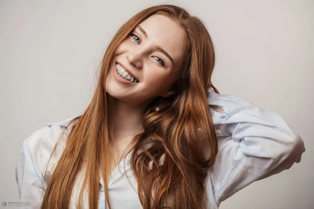 A smiling young woman with long red hair and braces at Redemption Orthodontics in Tallahassee, FL, poses against a plain background - Clear Braces vs Metal Braces in Tallahassee, FL