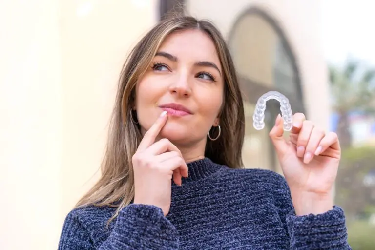 A woman in a grey sweater thoughtfully holds a clear dental aligner at Redemption Orthodontics in Tallahassee, FL - Aligners vs Braces in Tallahassee, FL