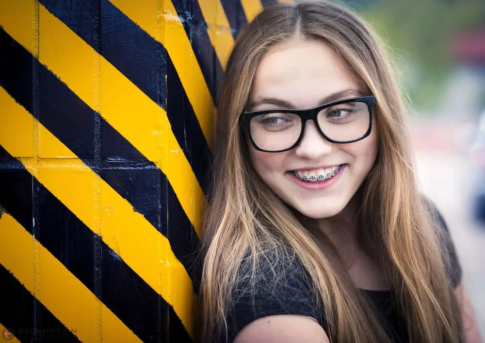 Smiling teen girl with braces from Redemption Orthodontics in Tallahassee, FL, leans on black and yellow striped wall - Aligners vs Braces in Tallahassee, FL
