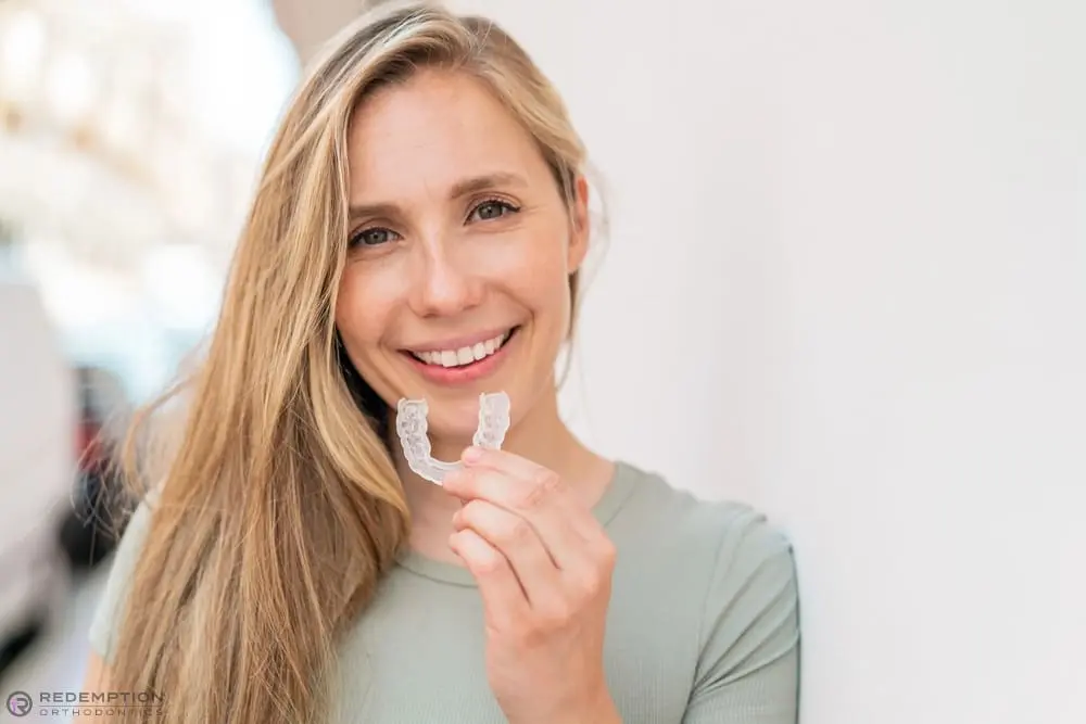 Smiling woman with long blonde hair holds a clear dental aligner at Redemption Orthodontics in Tallahassee, FL - Aligners vs Braces in Tallahassee, FL 