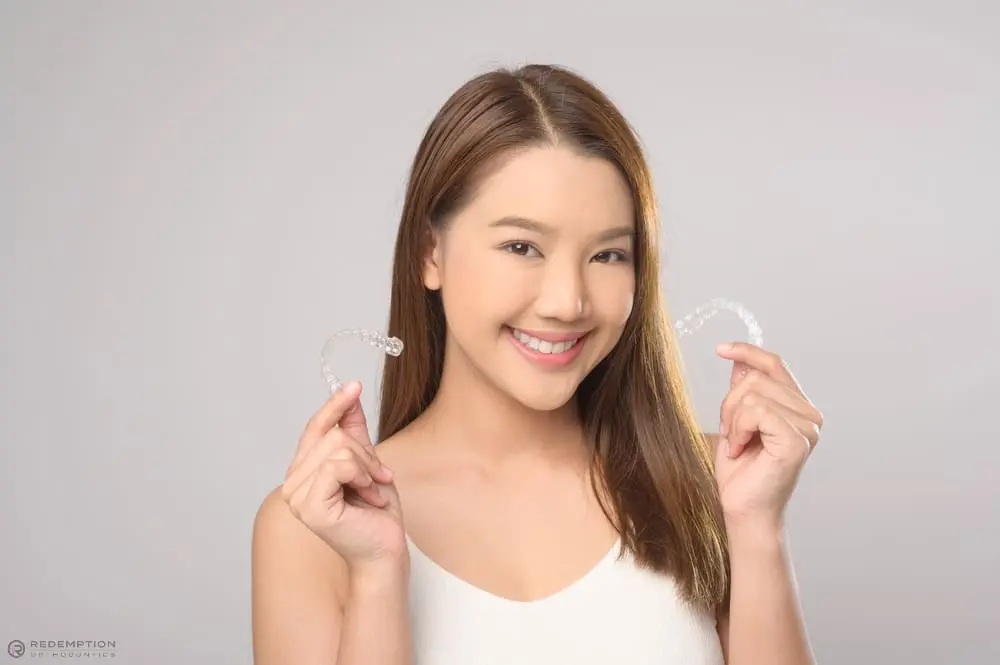A woman smiling holds up two clear dental aligners at Redemption Orthodontics in Tallahassee, FL, against a plain background - How Does Invisalign Work in Tallahassee, FL