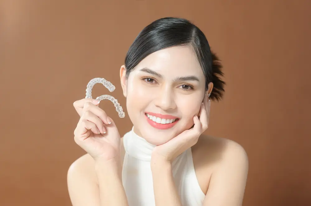 A woman at Redemption Orthodontics in Tallahassee, FL smiles holding two clear dental aligners against a plain brown background - How Does Invisalign Work in Tallahassee, FL