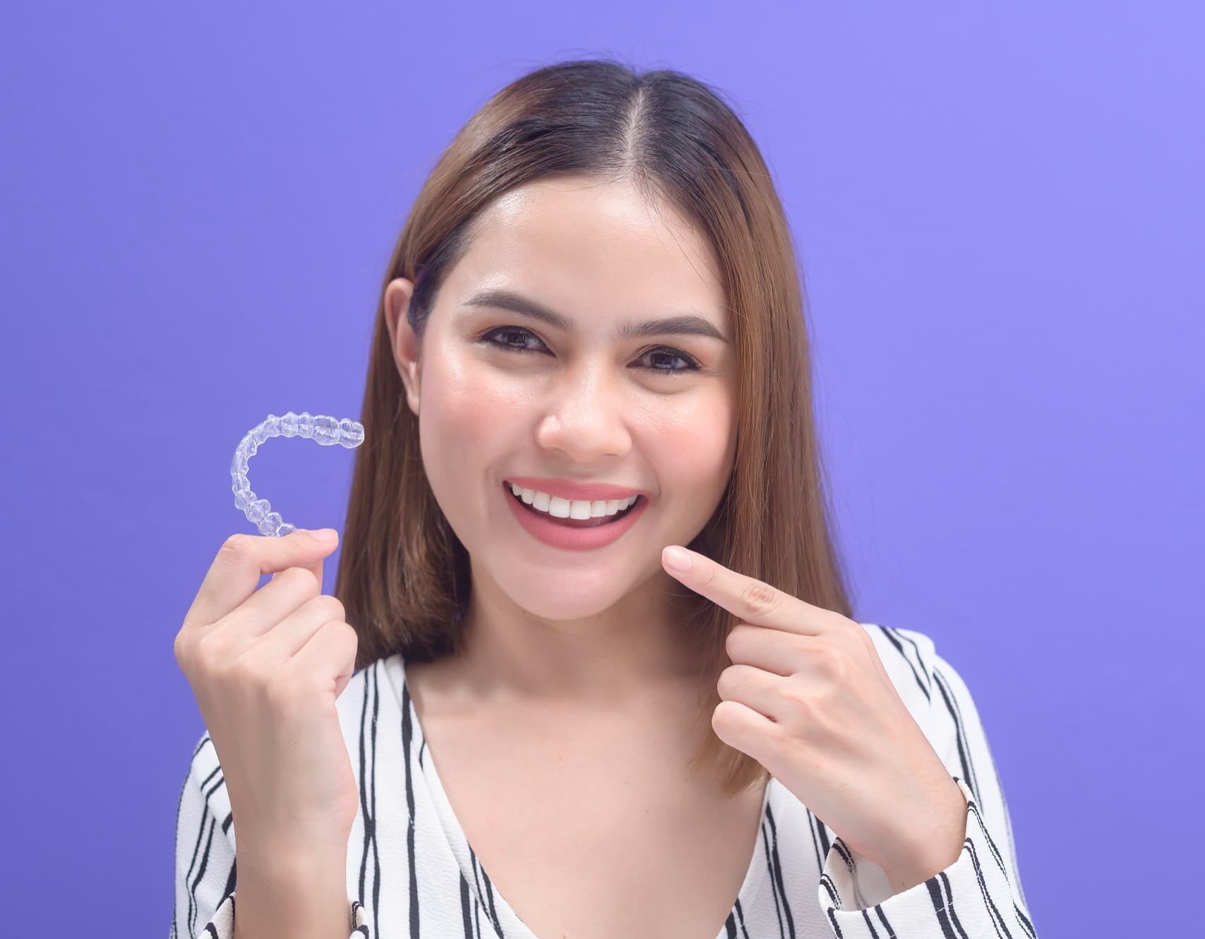 Smiling woman with clear dental aligner Invisalign points to her teeth, purple background; Redemption Orthodontics in Tallahassee, FL.